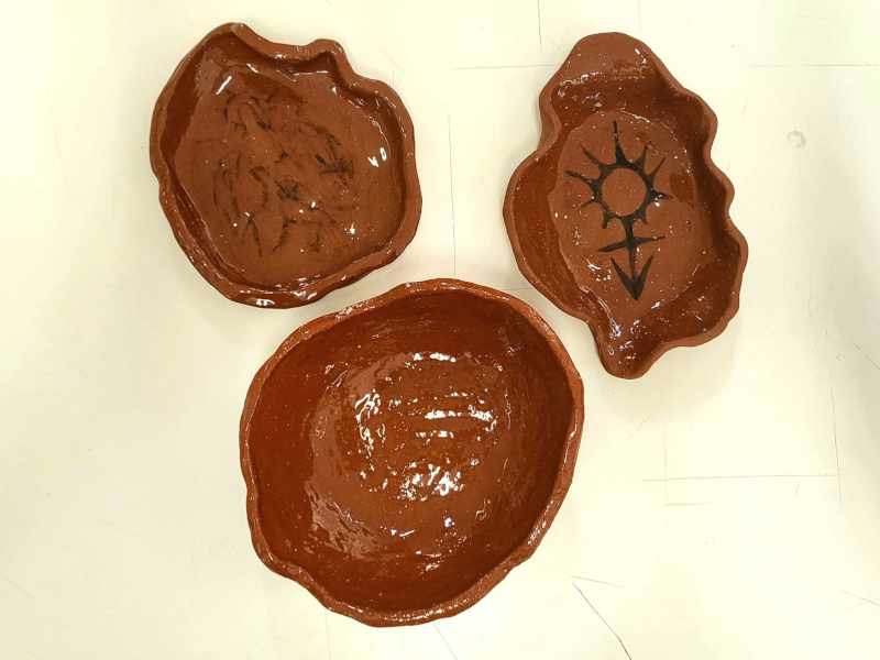 top view of three glazed, brick-colored pieces. one is a shalow, uneven dish with the faint drawing of a transgender angel. one is a deeper, leaf shaped dish with the mixed spoke of the transgender symbol. one is a simple round bowl